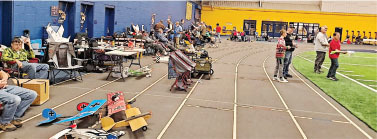 People in a gym setting, sitting and walking by tables with various items.