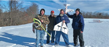 Four men on snowy field holding remote-controlled planes.