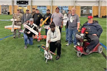 Group of people with model airplanes on indoor turf field.