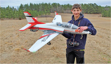 Young person smiling, holding a model jet plane outdoors in a field.