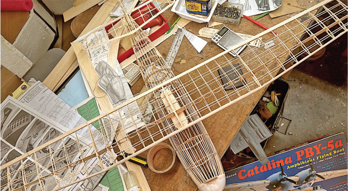 Model airplane framework on cluttered workbench with Catalina PBY-5a plans.