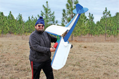 Man in a field holding a model airplane, wearing a winter hat and sunglasses.