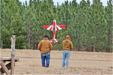 Two people watch a red and white model plane flying near trees.