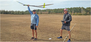 Two men in a field with a model glider, one holding a remote control.