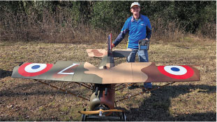 Man holding remote next to a large model biplane outdoors.
