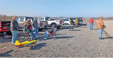People sitting and standing near trucks on gravel, with a yellow model plane in the foreground.