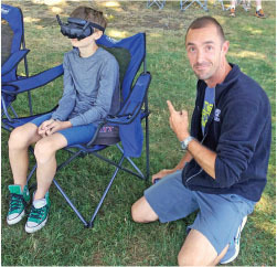 Child in VR headset sits on a chair, adult kneels next to them pointing.