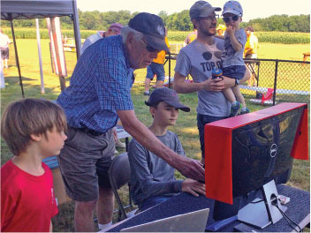 Group of people outdoors using a computer, with a child on an adult's shoulders.