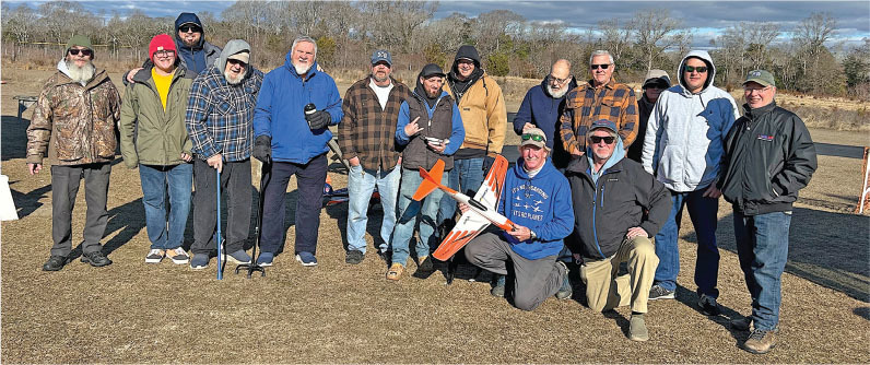 Group of 14 men outdoors, holding a model airplane. Cloudy sky and trees in the background.