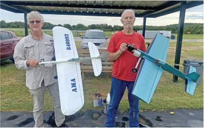 Two men holding model airplanes under a shelter at a grassy field.