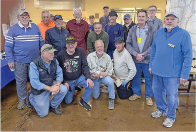 Group of men posing indoors, some seated, others standing, casual attire.