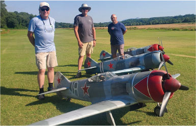 Three men stand beside model planes on a grassy field.