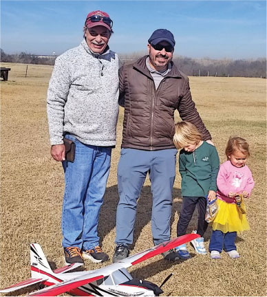 Two men and two children smiling in a field with a model airplane.