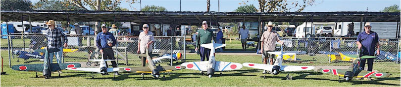 Seven men standing with model planes on grass, fence and pavilion in background.