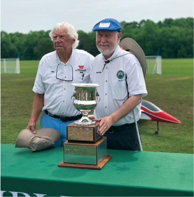 Two men with a trophy on a green table outdoors. One wears a blue cap.