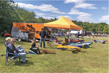 People sitting under an orange canopy with model planes on grass.