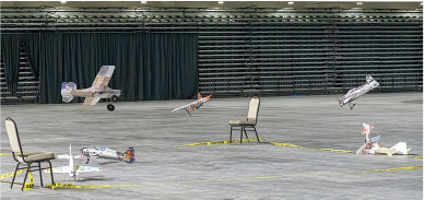 Remote-controlled planes flying indoors near empty chairs and a taped-off area.