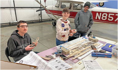 Three people at a table building a model airplane. A red and white plane in the background.