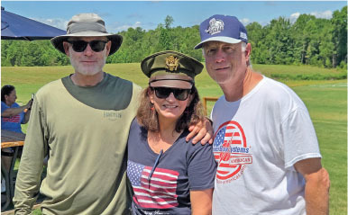 Three people outdoors, wearing hats and patriotic shirts, smiling in a grassy field.