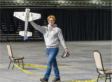 Person smiling, holding a model airplane and a controller in an indoor arena space.