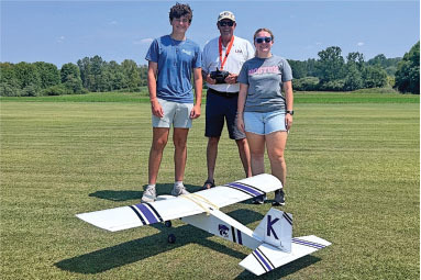 Three people stand on grass behind a model airplane, clear blue sky above.