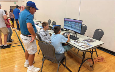 Two boys using a flight simulator, watched by a man in a gym.