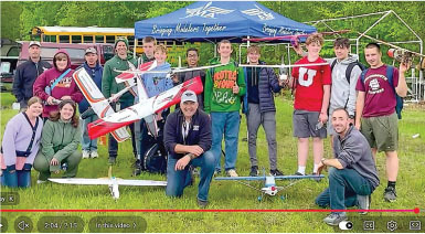 Group of people outdoors with model airplanes, blue tent behind them.