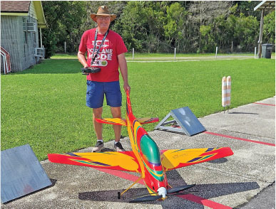 Man with red shirt holding remote, standing beside colorful model plane on grass.