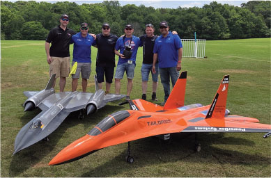 Group of six men standing behind two large model jets on grass.