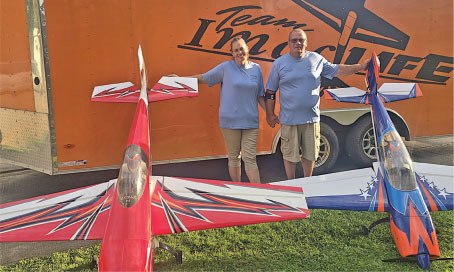 Two people in blue shirts beside large model planes and an orange trailer.
