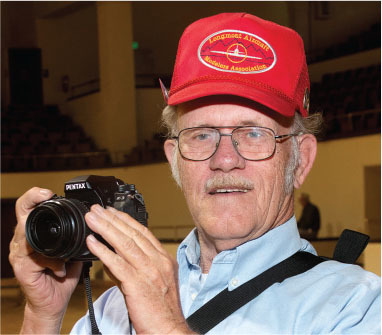 Man with glasses and red cap holding a Pentax camera indoors.