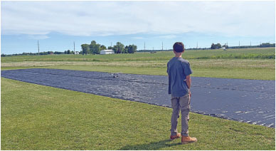 Boy standing on grass near large black tarp, under a blue sky.