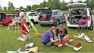 People assembling model airplanes next to open car trunks in a grassy area.