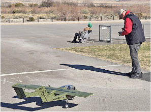 Man controlling a remote-controlled green plane on a runway. Another person sitting nearby.
