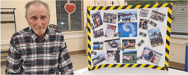 Elderly man standing beside a photo collage on a board with yellow caution tape border.