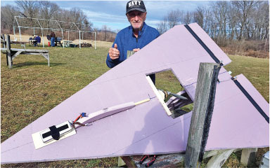 Man standing outdoors with a large purple model aircraft on a grassy field.