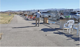 Man walking by model planes on tables near a fenced runway with mountains in the background.
