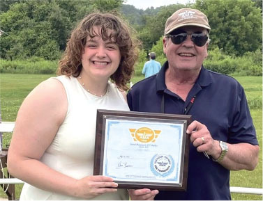 Man and woman smiling, holding a framed certificate outdoors.