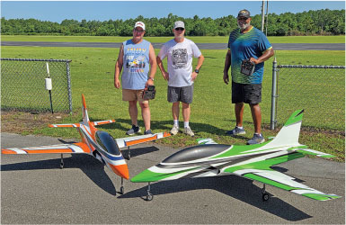 Three men with remote controls and two large model jet planes on an airstrip.