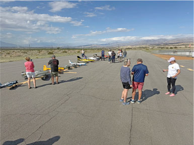 People standing near remote-controlled airplanes on a sunny runway.