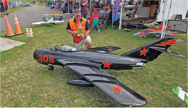 Man kneeling beside large model jet with red stars and number 309 on grassy field.
