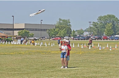 People flying model aircraft in a grassy field, with cones and parked cars in the background.