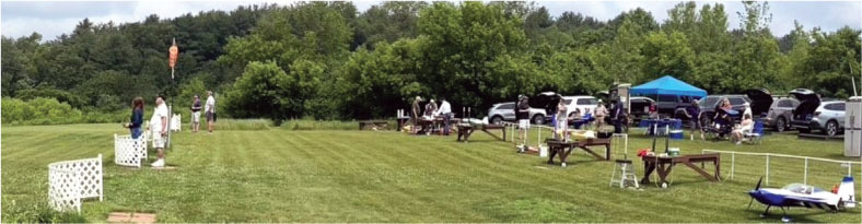 People at a grassy field set up with tables and cars, surrounded by trees.