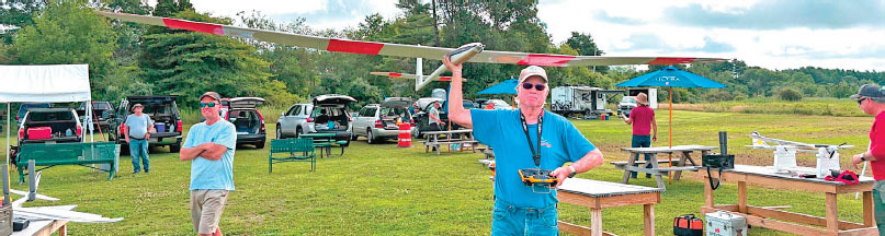 Man holding a large model glider at a field with parked cars and trees in the background.