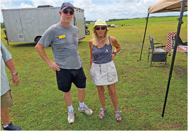 A man and woman stand on grass near a trailer and canopy, under a partly cloudy sky.