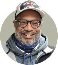 Smiling man wearing glasses and a cap with a blue scarf and gray hoodie.