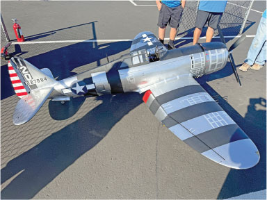 Silver model aircraft with black stripes on pavement, people in background.