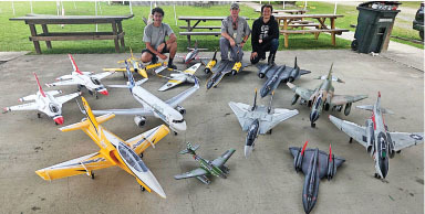 Three people seated behind an array of model jets on concrete.
