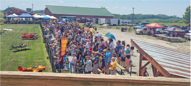 Crowd gathered outdoors near a fence, with tents and small airplanes nearby.