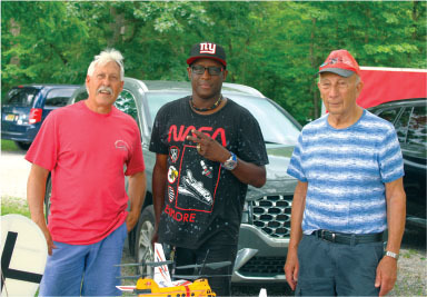 Three men standing in front of parked cars, one wearing a NASA T-shirt and cap.